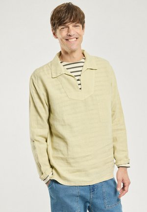 Homme souriant debout, les mains dans les poches, portant une chemise à manches longues beige clair sur un haut rayé et un jean bleu devant un fond uni.