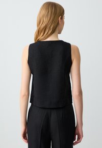 Top nero senza maniche realizzato in tessuto texturizzato, con collo alto e linea dritta. Indossato con pantaloni neri abbinati.