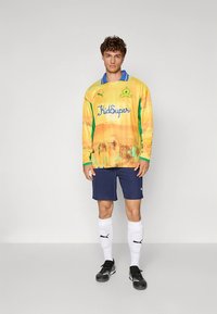 Maglia da calcio a maniche lunghe multicolore con sfumatura gialla, dettagli verdi e colletto blu, abbinata a pantaloncini blu scuro e calzini bianchi.