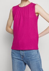 Haut sans manches rose vif texturé avec petite bordure de perles sur le col et les emmanchures, porté avec un jean denim bleu clair.
