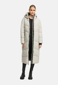 Donna che indossa un lungo piumino beige chiaro con cappuccio, top nero, jeans neri e stivaletti neri, in piedi su uno sfondo bianco.