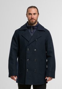 Marineblauer, doppelt geknöpfter Wollmantel mit großen Knöpfen, weitem Kragen und seitlichen Taschen; über einer dunklen, gesteppten Innjacke getragen.