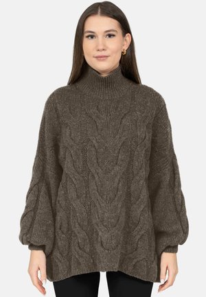 Frau trägt einen braunen, übergroßen Rollkragenpullover mit Kabelstrickmuster und Ballonärmeln, sie steht vor einem einfarbigen Hintergrund.