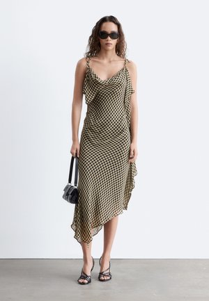 Femme portant une robe asymétrique à carreaux noirs et beige avec des bretelles spaghetti, des talons noirs, des lunettes de soleil, et portant un petit sac à main noir.