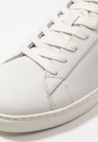 Zapatilla de piel blanca con cordones planos blancos, punta redonda y costuras sutiles. Textura suave con suela de goma y diseño minimalista.