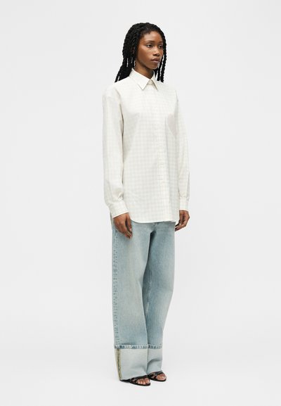 Γυναίκα με μακριά κοτσίδα που φοράει oversized λευκό πουκάμισο με γιακά, ανοιχτόχρωμα τζιν με φαρδιές παντελόνες και μεγάλες μανσέτες, και μαύρες ανοιχτές γόβες.