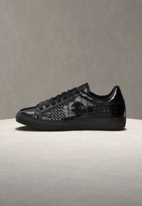 Baskets basses noires en cuir texturé et brillant, à lacets sur le devant, avec un logo embossé sur le côté, présentées sur une surface neutre.