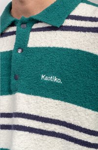 Svītrots polo krekls gaiši zilā, baltā un tumši zilā krāsā, izgatavots no teksturēta auduma. Tam ir apkakle un pogu aizdare, ar "Kaotiko." logotipu.