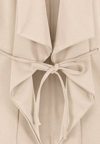 Ceinture en tissu beige nouée en un nœud autour d'un vêtement drapé à texture douce, avec de larges revers pointus.