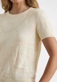 Krémový sveter s krátkym rukávom, štruktúrovaným pleteným vzorom, okrúhlym výstrihom a rebrovanými manžetami a lemom. Vyrobený z ľahkej látky.