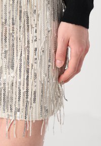 Main reposant à côté d'une jupe beige ornée de rangées verticales de sequins argentés et noirs, sur un fond clair uni.