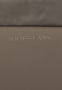 Hnedá textúrovaná látka s hladkým hnedým panelom nižšie, s vyrazeným logom "JOOP". Dizajn je minimalistický a elegantný.