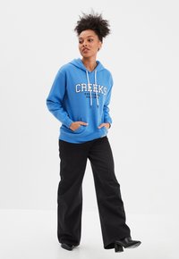 Sudadera azul con impresión frontal, capucha con cordones y bolsillo tipo canguro, combinada con pantalones de pierna ancha negros y botines de tacón. Textura de tela suave.