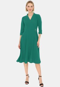 Zielona sukienka kopertowa z rękawami o długości trzech ćwiartek, dekoltem w kształcie litery V, dopasowanym w pasie, rozkloszowanym dołem oraz akcentami w kolorze srebrnym. Gładka tekstura materiału.