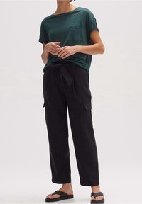 T-shirt vert foncé à manches courtes avec une poche poitrine, associée à un pantalon cargo noir taille haute avec ceinture nouée et poches latérales.