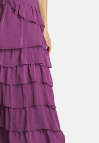 Lila gestufte Kleid mit Rüschenschichten, aus einem strukturierten Stoff und asymmetrischem Saum. Nahaufnahme einer Hand auf dem Stoff.