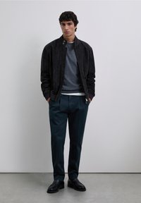 Svart bombarjacka i mocka, grå tröja, blågröna byxor och svarta skor. Modellen står i en neutral miljö med händerna i fickorna.