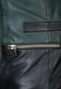 Černá a zelená kožená bunda s dekorativní černou nášivkou a zlatým zipem na dolní levé straně, která ukazuje texturovaný materiál.