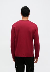 Koszula z długim rękawem w ciemnoczerwonym kolorze, wykonana z bawełnianej tkaniny, z okrągłym dekoltem, luźnym krojem i długością sięgającą do bioder, o minimalistycznym designie.