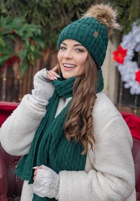 Gorro de punto verde con un pompón de piel, bufanda de punto a juego y guantes sin dedos grises. Lleva puesto un abrigo de piel blanco por encima.