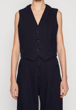 Mouwloos donkerblauw vest met drie knopen aan de voorkant, gedragen met bijpassende broek met hoge taille en zijknopdetails.