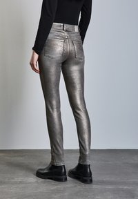 Metallisk grå skinny jeans med hög midja, fem fickor och en slät yta, tillsammans med svarta ankelskodlor på en grå bakgrund.