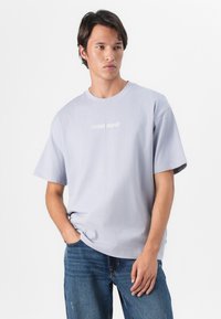 Světle levandulové bavlněné tričko s krátkým rukávem a kulatým výstřihem, na kterém je po celé šířce hrudi bílé logo "GUESS JEANS".