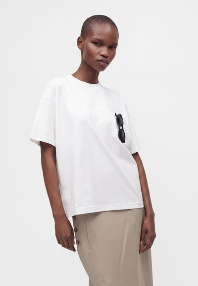 Γυναίκα με κοντά μαλλιά που φοράει oversized λευκό μπλουζάκι με γυαλιά ηλίου στην τσέπη του στήθους και μπεζ φούστα με κουμπιά στο πλάι.
