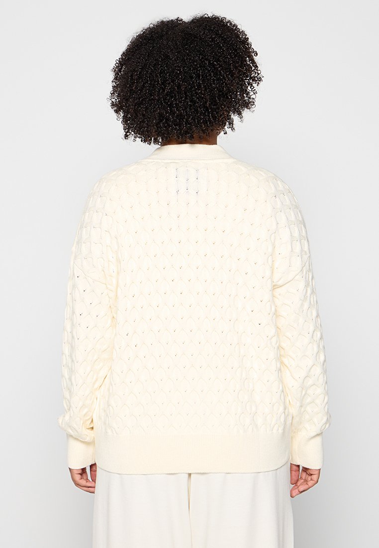 Crème-kleurige gebreide trui met een getextureerd ruitpatroon, geribde manchetten en een relaxed fit. De achterkant toont gedetailleerde stiksels.