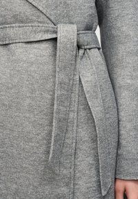 Ύφασμα γκρι μάλλινου παλτού με ταιριαστή ζώνη δεμένη σε χαλαρό κόμπο στη μέση, που δείχνει μέρος ενός μανικιού και χέρι.