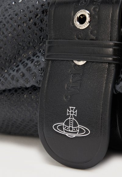 Μαύρο δερμάτινο λουράκι σανδαλιού με ασημένιες οπές και ανάγλυφη επωνυμία, τοποθετημένο σε φόντο με μαύρο ύφασμα με σχέδιο.