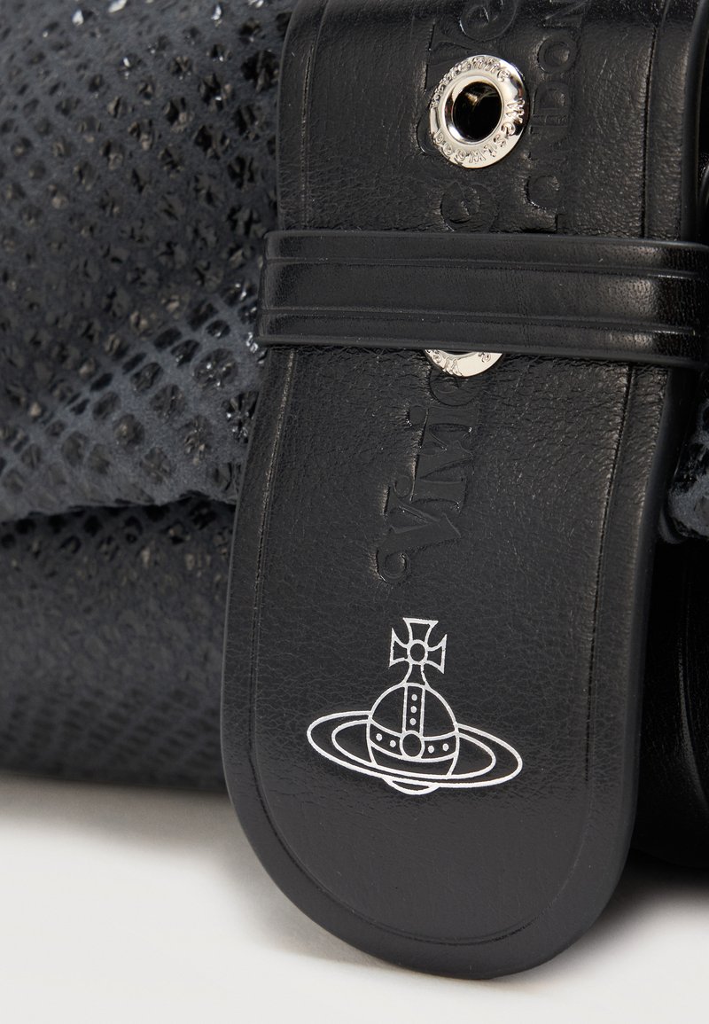 Bride de sandale en cuir noir avec œillets argentés et nom de marque embossé, posée sur un fond de tissu noir à motif.