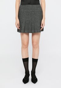 Grå plissert miniskjørt med fiskebensmønster, ledsaget av svarte gjennomsiktige sokker og spisse svarte sko, mot en ensfarget hvit bakgrunn.