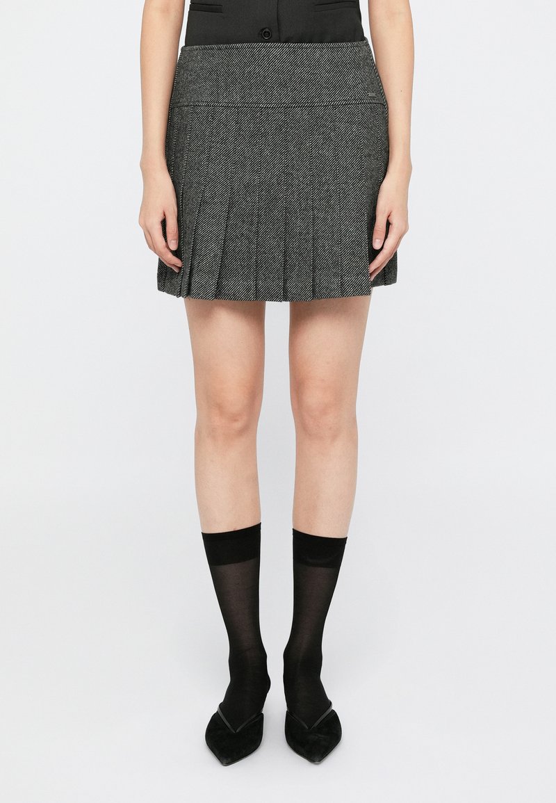 Grå plissert miniskjørt med fiskebensmønster, ledsaget av svarte gjennomsiktige sokker og spisse svarte sko, mot en ensfarget hvit bakgrunn.