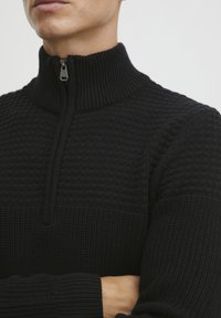 Pull en tricot noir avec col montant, fermeture éclair à mi-hauteur et motif texturé sur la poitrine. Manches et ourlet côtelés.