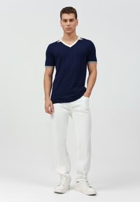 Navyblauer kurzärmeliger V-Ausschnitt-Pullover mit weißen gestreiften Akzenten am Kragen und an den Ärmeln, kombiniert mit weißen Hosen und weißen Sneakers.