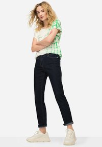 Camiseta blanca con diseño de cuadros verdes, vaqueros oscuros y zapatillas ligeras. El conjunto presenta un corte relajado y los puños de los vaqueros están enrollados.