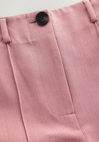 Pantalon rose sur mesure avec un tissu texturé, des poches latérales et un détail de bouton noir unique à la taille.
