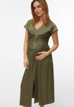 Mujer embarazada con cabello hasta los hombros, vestida con un vestido midi verde oliva, sosteniendo suavemente su vientre contra un fondo liso.