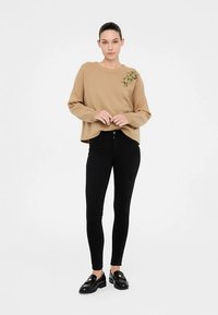 Maglione beige lavorato a maglia con trama a coste, caratterizzato da un ornamento floreale multicolore sulla spalla sinistra, abbinato a jeans skinny neri e mocassini.