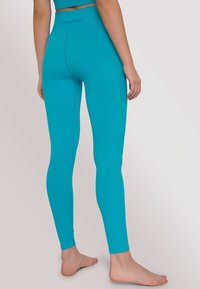 Donna in piedi a piedi nudi, che indossa leggings a vita alta color turchese e un top corto abbinato, girata di spalle su uno sfondo neutro.