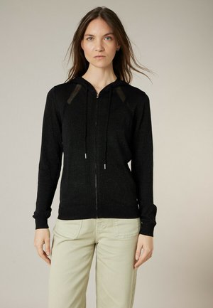 Femme portant un sweat à capuche noir à fermeture éclair et un pantalon beige clair, debout devant un fond neutre uni.