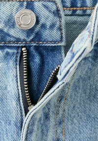 Detailní záběr světle modrých džínů z denimu s částečně otevřeným stříbrným zipem a kovovým knoflíkem s nápisem "Jeans Established 1984."