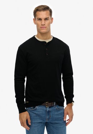 Mężczyzna ubrany w czarną koszulę z długim rękawem na guziki, na beżowej podkoszulce, niebieskie dżinsy, brązowy pasek i złoty zegarek na rękę, stojący na białym tle.