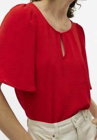 Rød bluse lavet af struktureret stof, med en nøglehulåbning ved halsen og pufærmer. Bæres med beige højtaljede bukser.