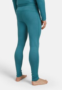 Persona che indossa leggings aderenti color turchese e una maglietta a maniche lunghe coordinata, in piedi a piedi nudi su uno sfondo bianco, mostrata di spalle.
