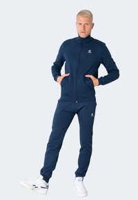 Chándal azul marino de tejido suave, con chaqueta de cremallera y pantalones a juego con bolsillos. Incluye detalle de logo en el pecho.