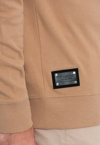 Beige Sweatshirt mit gerippten Bündchen, versehen mit einem schwarzen Leder-Patch mit silbernen Akzenten. Glatte Textur und schlichtes Design.
