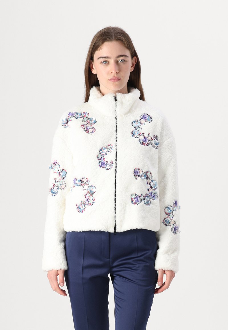 Weiße Kunstpelzjacke mit floralen Stickereien in Blau und Violett, mit einem Reißverschluss vorne und einem hohen Kragen, getragen über navyblauen Hosen.