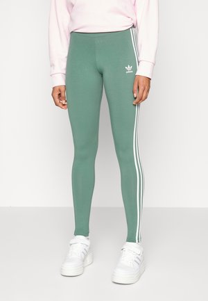 Leggings verdes hechos de una tela suave con un cinturón alto, que presentan tres franjas laterales blancas y un logo de Adidas en la cadera izquierda.